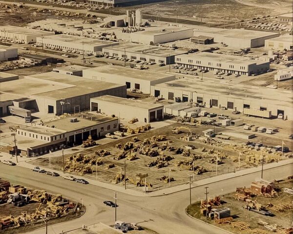 Krysa lot A vintage aerial photograph of the original industrial lot and warehouse facility, showing numerous pieces of heavy machinery and construction equipment parked in a central yard.